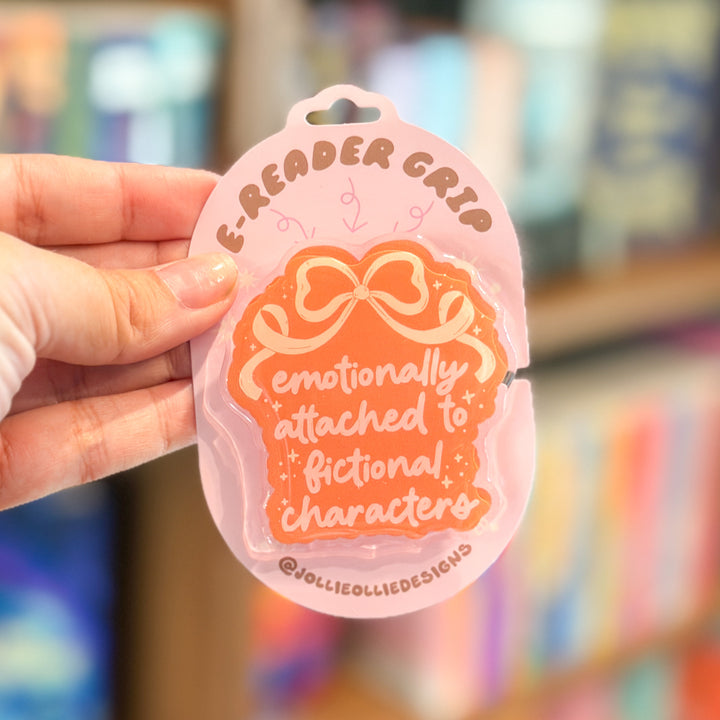 A hand holds a pink and orange sticker shaped like a book cover with the text &