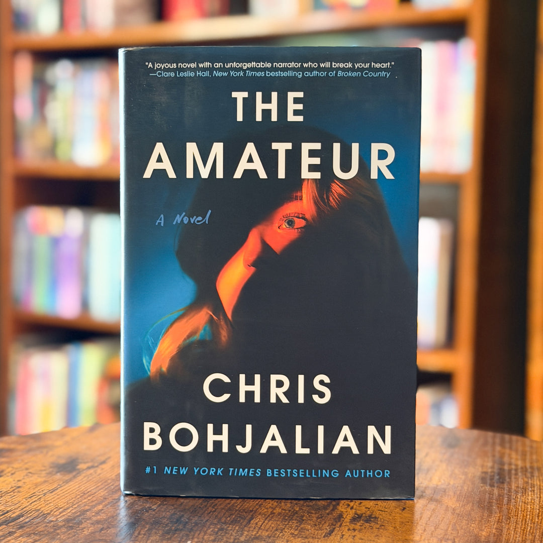 The book cover of 'The Amateur' by Chris Bohjalian, standing upright on a wooden table with blurred bookshelves in the background.