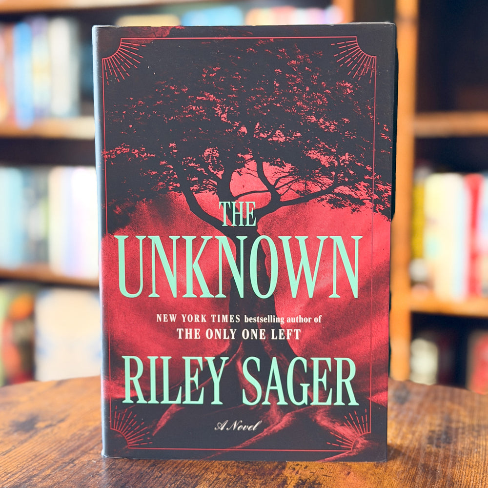 The book cover of 'The Unknown' by Riley Sager, featuring a dark silhouette of a tree against a vibrant red and pink sky, displayed on a wooden table with blurred bookshelves in the background.
