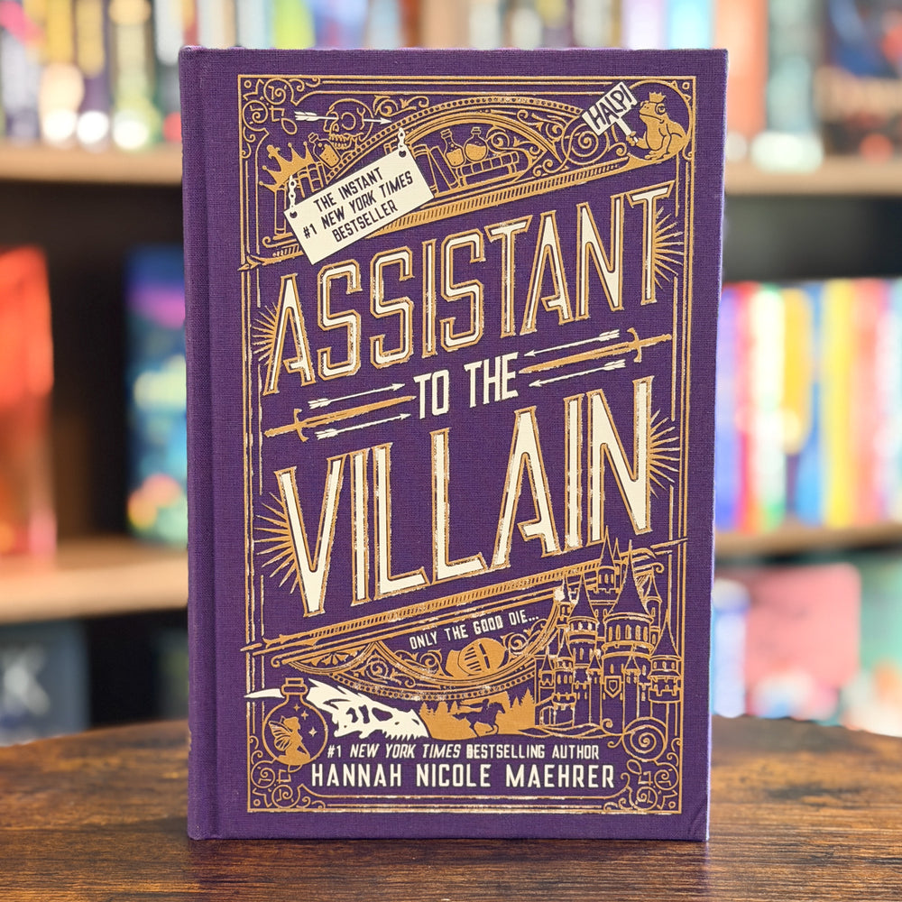 The book 'Assistant to the Villain' by Hannah Nicole Maehrer, featuring a purple cover with ornate gold illustrations and lettering, standing on a wooden table with a blurred bookstore background.