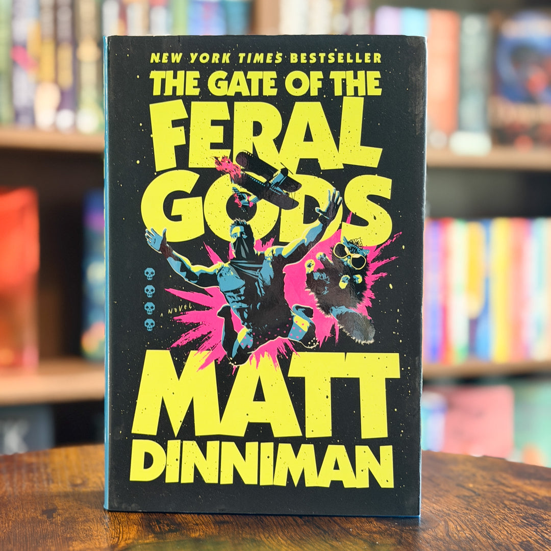The Gate of the Feral Gods book by Matt Dinniman, standing upright on a wooden table with a blurred bookshelf background.