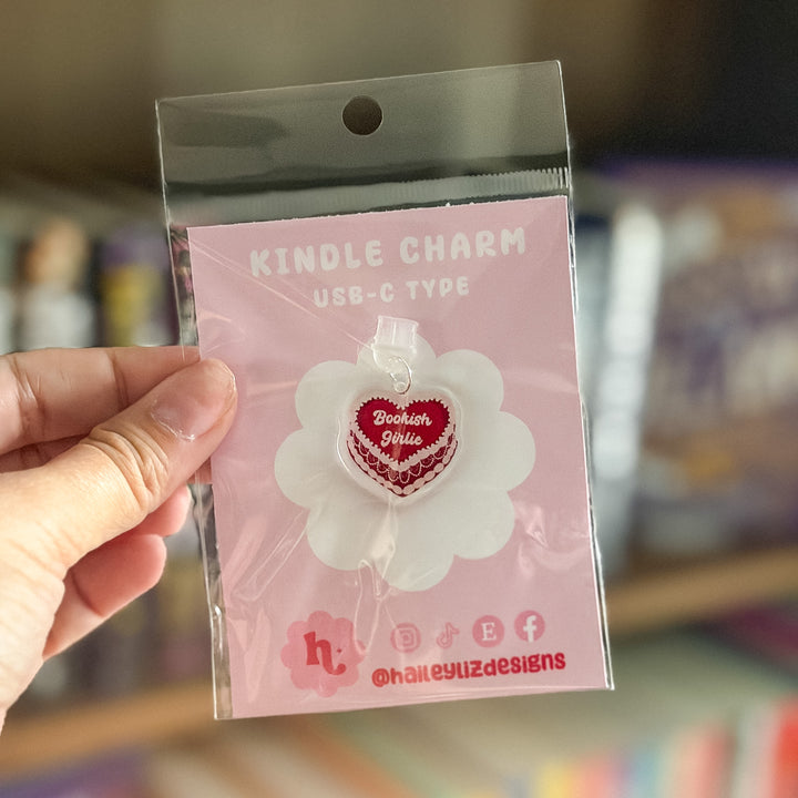 A hand holding a clear plastic blister pack containing a red heart-shaped USB-C charging charm with the text &