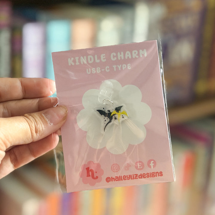 A hand holding a pink card displaying a small USB-C type Kindle charm shaped like a black and white dragon with yellow accents, set against a blurred bookshelf background.