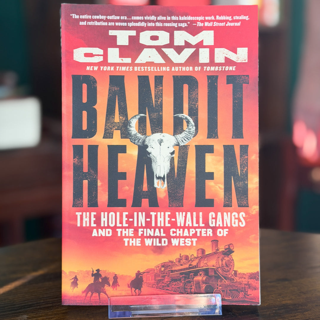 A copy of the book 'Bandit Heaven' by Tom Clavin standing upright on a clear plastic display stand on a wooden table. The book cover features a vibrant red and orange sunset background with a central black buffalo skull graphic and silhouettes of bandits on horseback and a train.