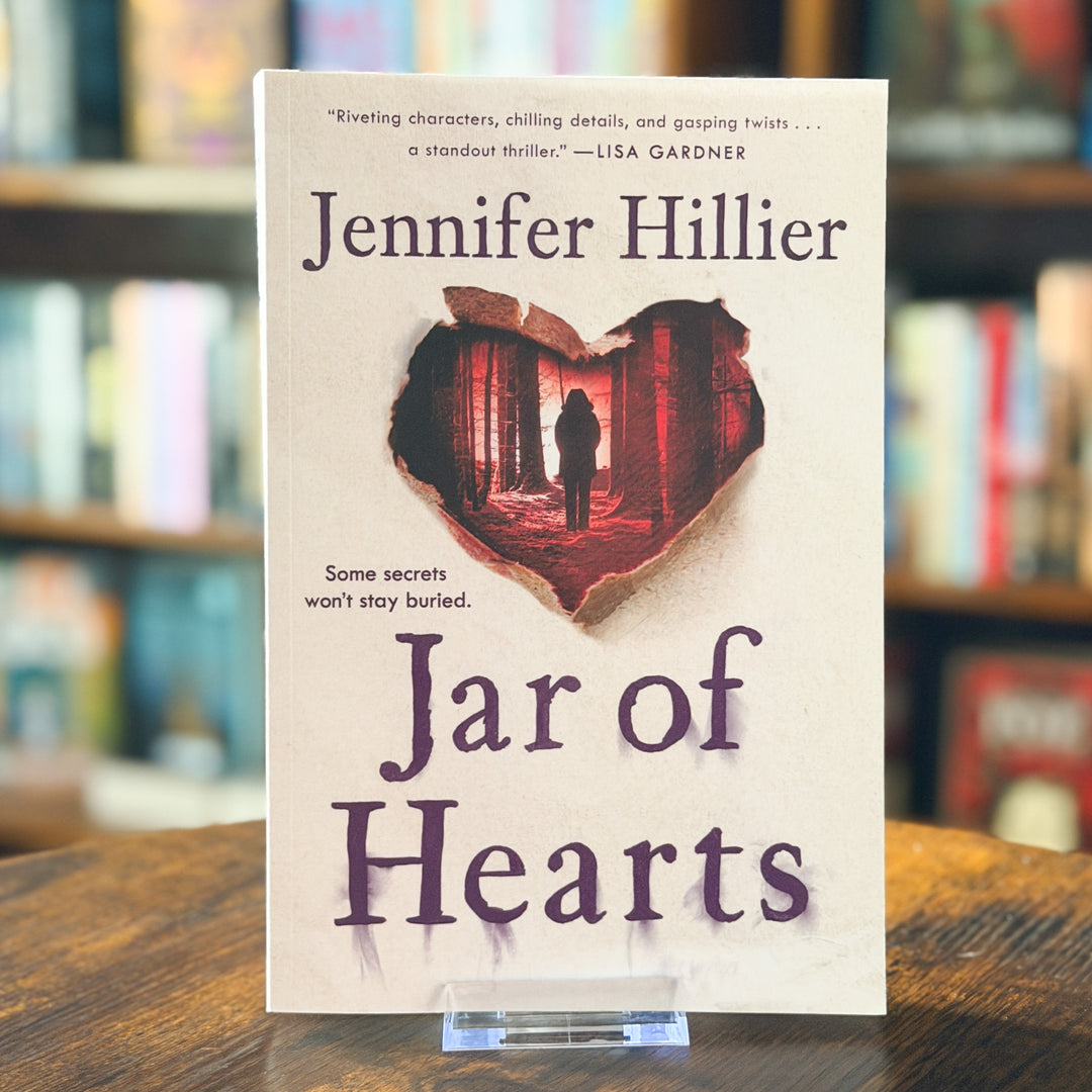 Book cover for 'Jar of Hearts' by Jennifer Hillier, displayed upright on a small clear stand on a wooden table with blurred bookshelves in the background.