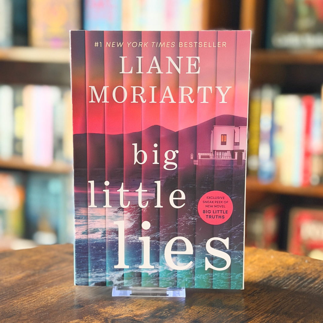 The book 'Big Little Lies' by Liane Moriarty stands upright on a clear plastic display stand on a wooden table, with a blurred bookshelf background.