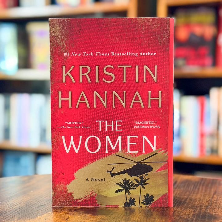 The Women novel by Kristin Hannah standing upright on a wooden table with blurred bookshelves in the background. The book cover is predominantly red with gold textured details, white title text, and a gold-toned illustration of a helicopter and palm trees near the bottom.