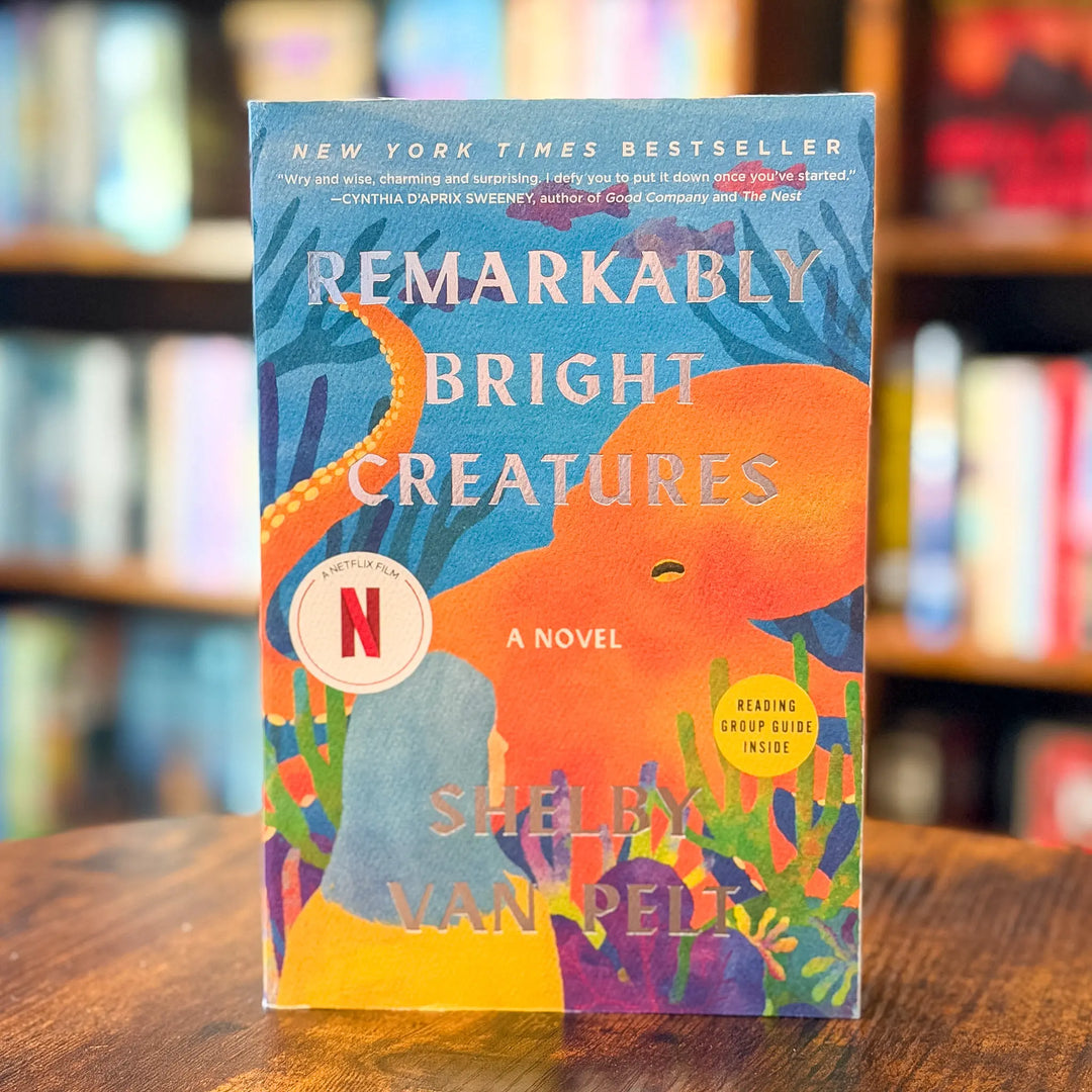 The book 'Remarkably Bright Creatures' by Shelby Van Pelt stands upright on a wooden table with a blurred library or bookstore background. The cover features a vibrant, colorful illustration of an octopus and coral reef, with the title in large white letters and a prominent circular Netflix logo badge.