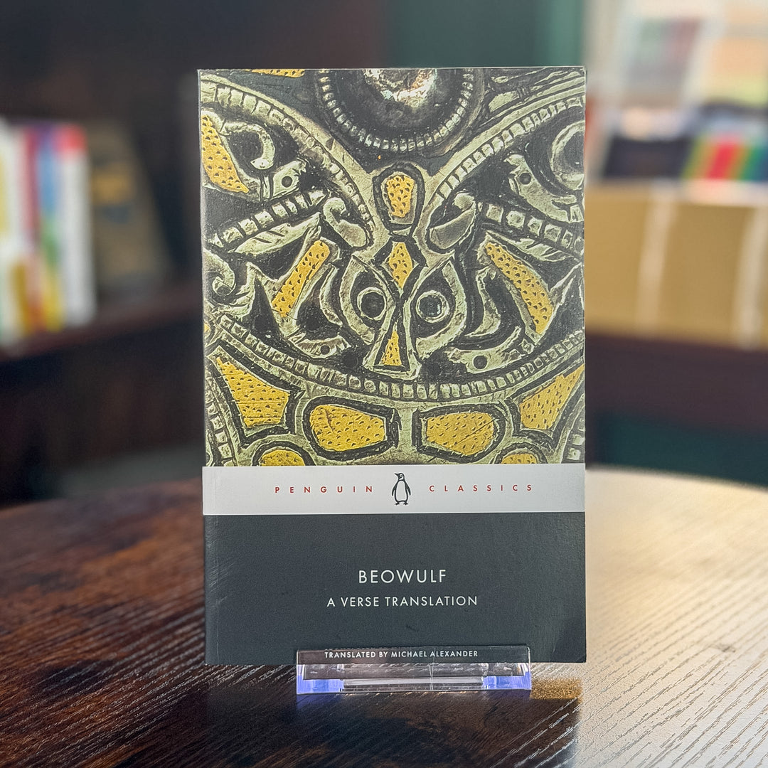 Beowulf: A Verse Translation paperback book by Penguin Classics, featuring a detailed black and gold ancient artifact illustration on the cover, displayed on a clear acrylic stand on a wooden table.