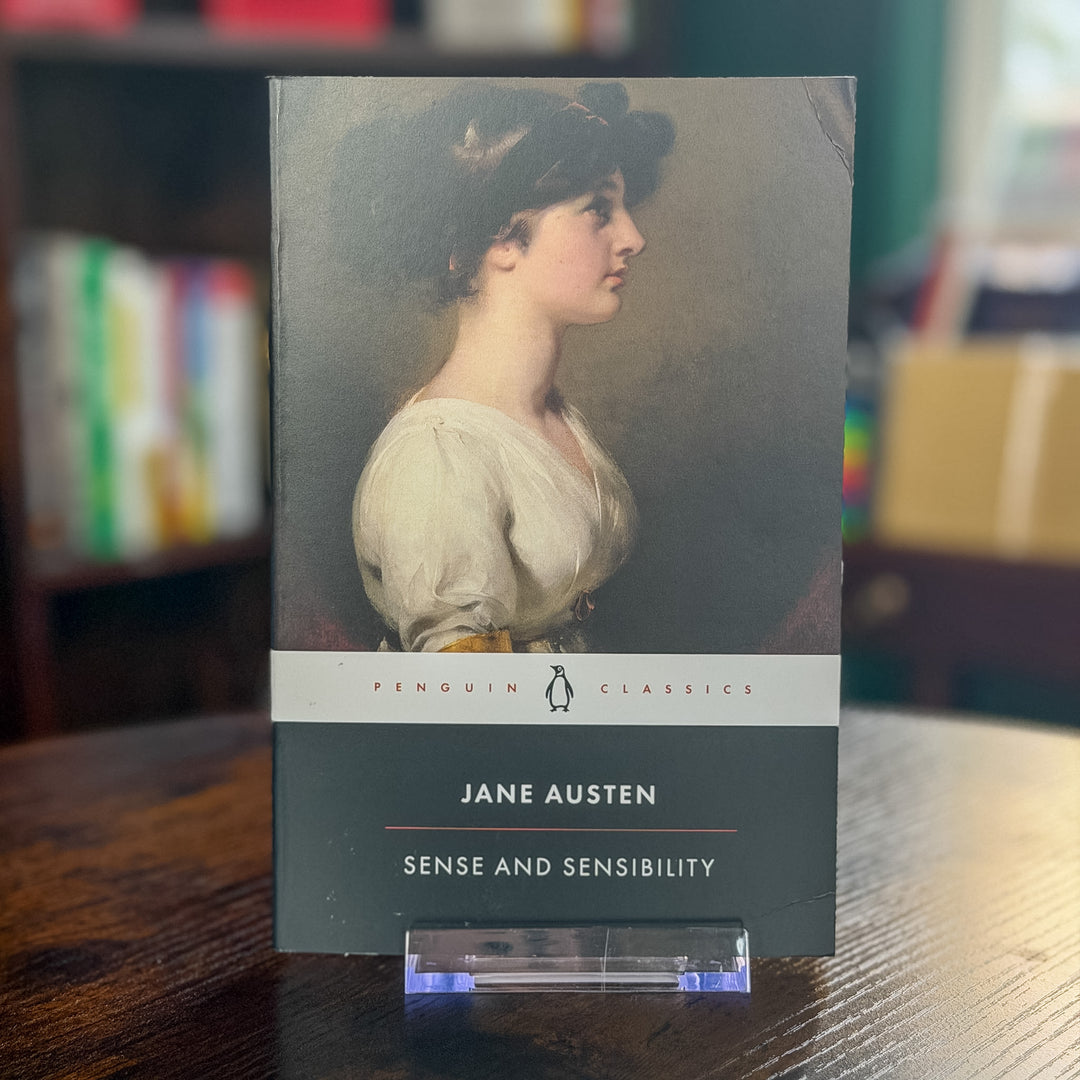 A paperback edition of Jane Austen's 'Sense and Sensibility' by Penguin Classics, displayed on a clear acrylic stand on a dark wooden table with a blurred bookshelf in the background.