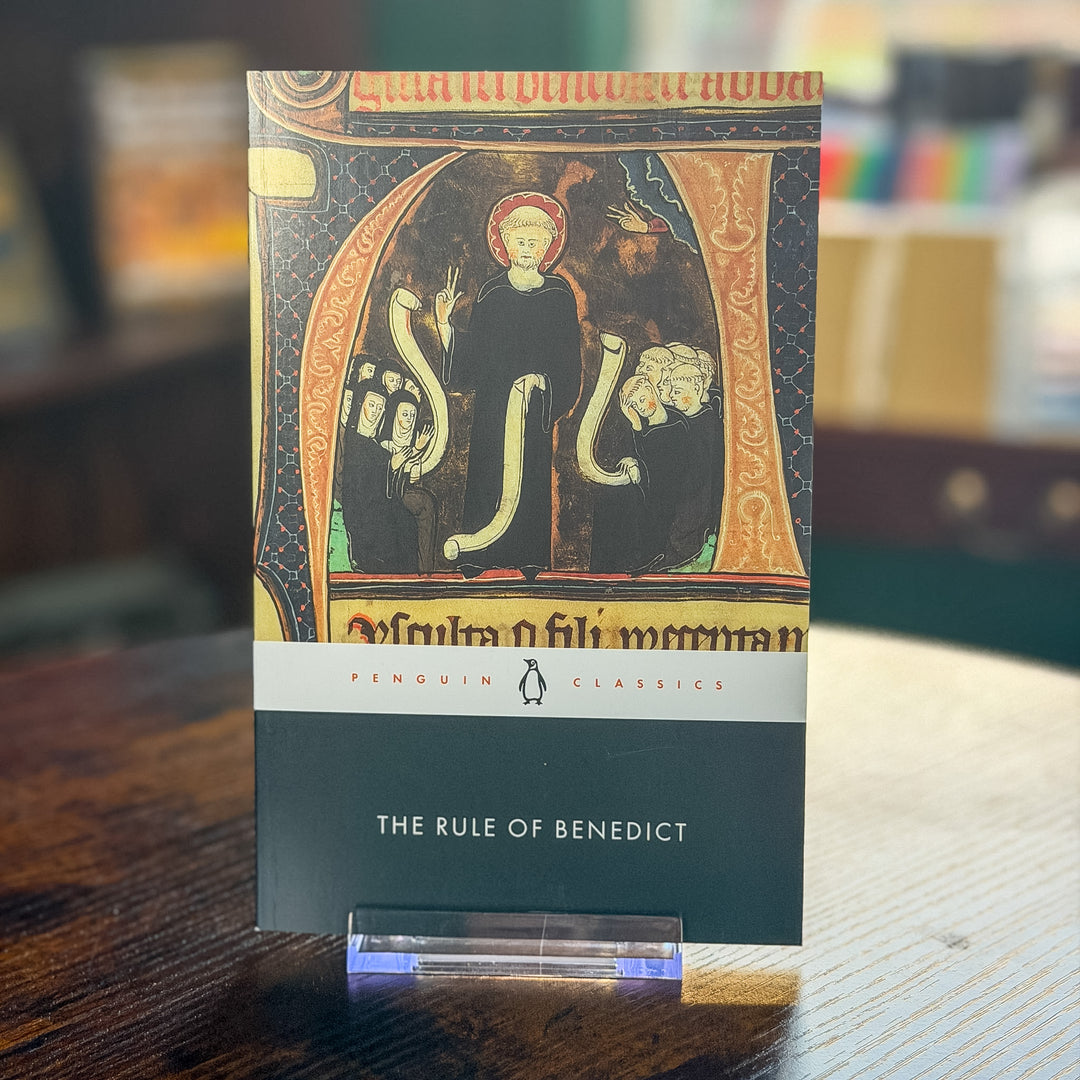 A book titled "The Rule of Benedict" by Penguin Classics stands upright on a small clear acrylic display block on a wooden table. The cover features a medieval-style illustration with a robed figure and scroll-like elements, with the title and Penguin logo visible.