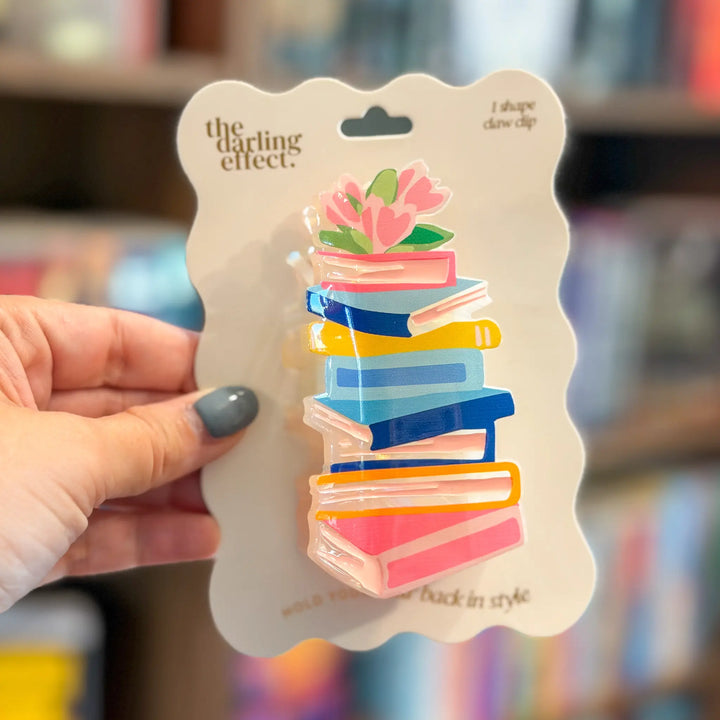 A hand holds a wavy-edged card displaying a book-stack claw clip. The clip features stacked books in pink, blue, yellow, and white with a pink flower on top.