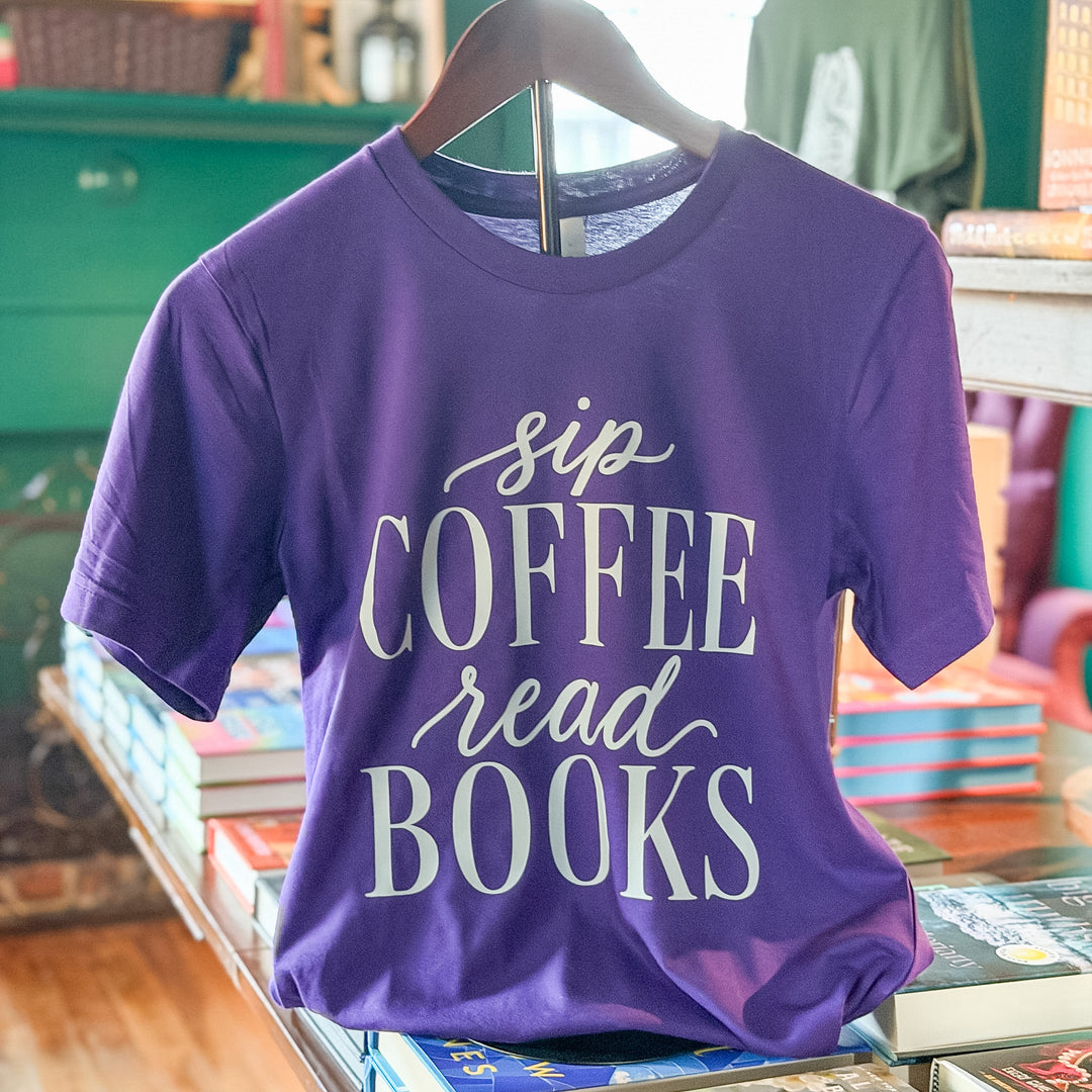 A purple short-sleeve graphic T-shirt hanging on a wooden hanger, with the phrase 'sip COFFEE read BOOKS' printed in white script and block lettering. The shirt is displayed on a bookshelf in a retail or bookstore setting with books visible in the background.