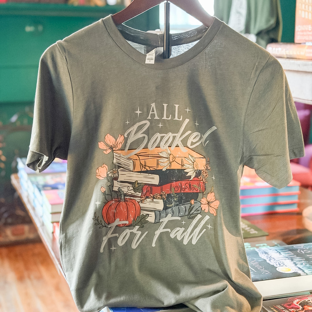A short-sleeve graphic t-shirt in a muted sage green color, hanging on a wooden hanger. The shirt features a fall-themed graphic with the text 'ALL Booked For Fall' in white script, surrounded by illustrations of a stack of books, a small orange pumpkin, and pink flowers.