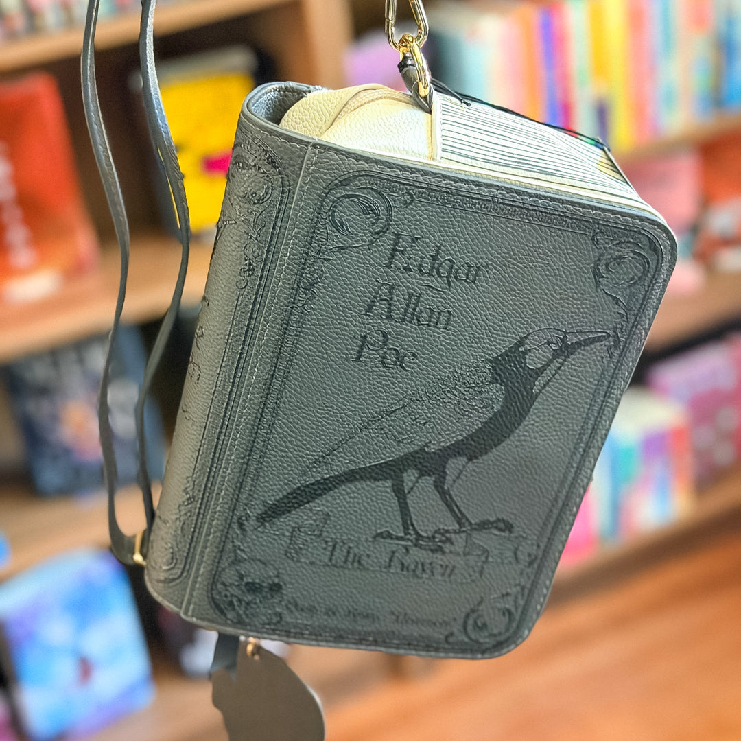 A book-shaped crossbody bag featuring Edgar Allan Poe's 'The Raven' design, hanging in front of a blurred bookstore background.