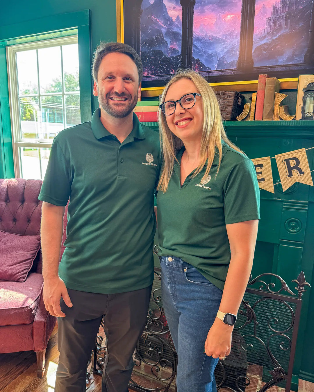 Two people in matching green polos standing in a room with a colorful painting and window.