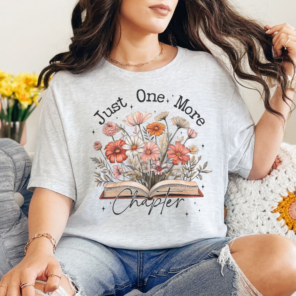 Person wearing a gray t-shirt with floral design and text 'Just One More Chapter'.