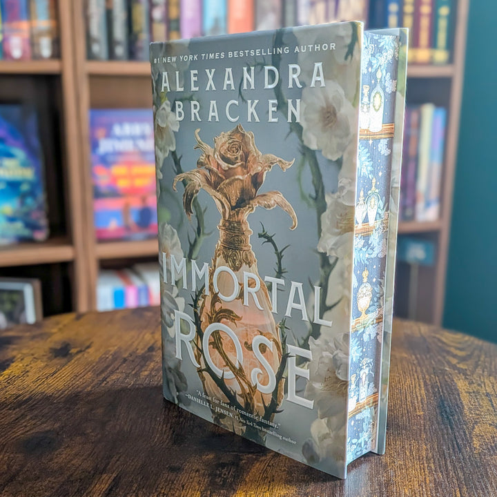 A hardcover book titled "Immortal Rose" by Alexandra Bracken, standing upright on a wooden table with a blurred bookshelf background.