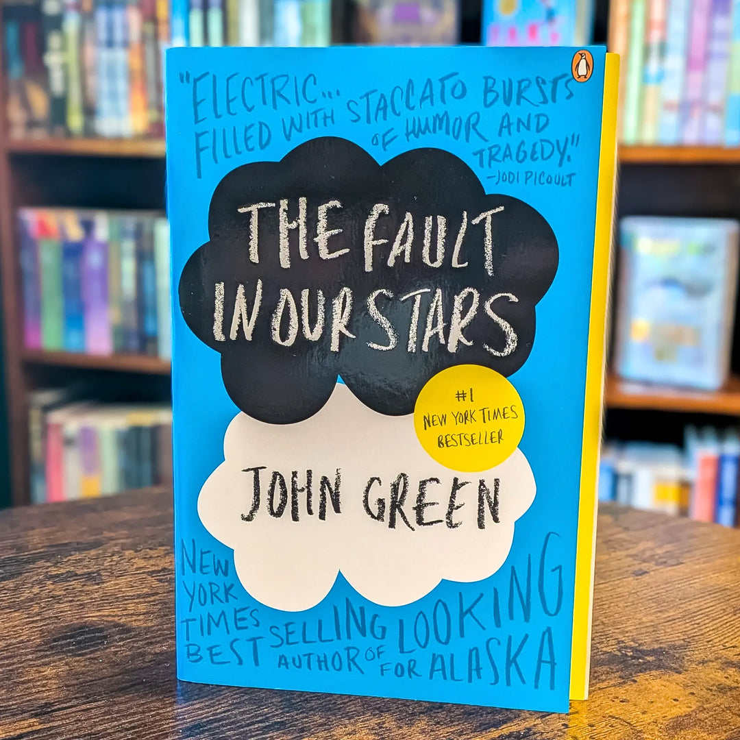 The Fault in Our Stars book by John Green, standing upright on a wooden table with blurred bookshelves in the background. The cover is bright blue with black and white cloud shapes containing the title and author name, plus a yellow circle badge.