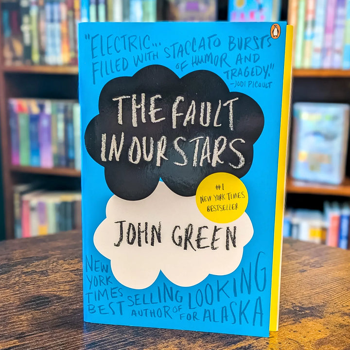 The Fault in Our Stars book by John Green, standing upright on a wooden table with blurred bookshelves in the background. The cover is bright blue with black and white cloud shapes containing the title and author name, plus a yellow circle badge.