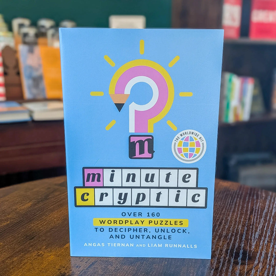 The front cover of the book 'The Minute Cryptic' stands upright on a wooden table, with a blurred library or study background showing books and shelves. The cover is light blue with a large pink and yellow question mark graphic, colorful block text reading 'minute cryptic,' and a small 'The World's Best' badge.