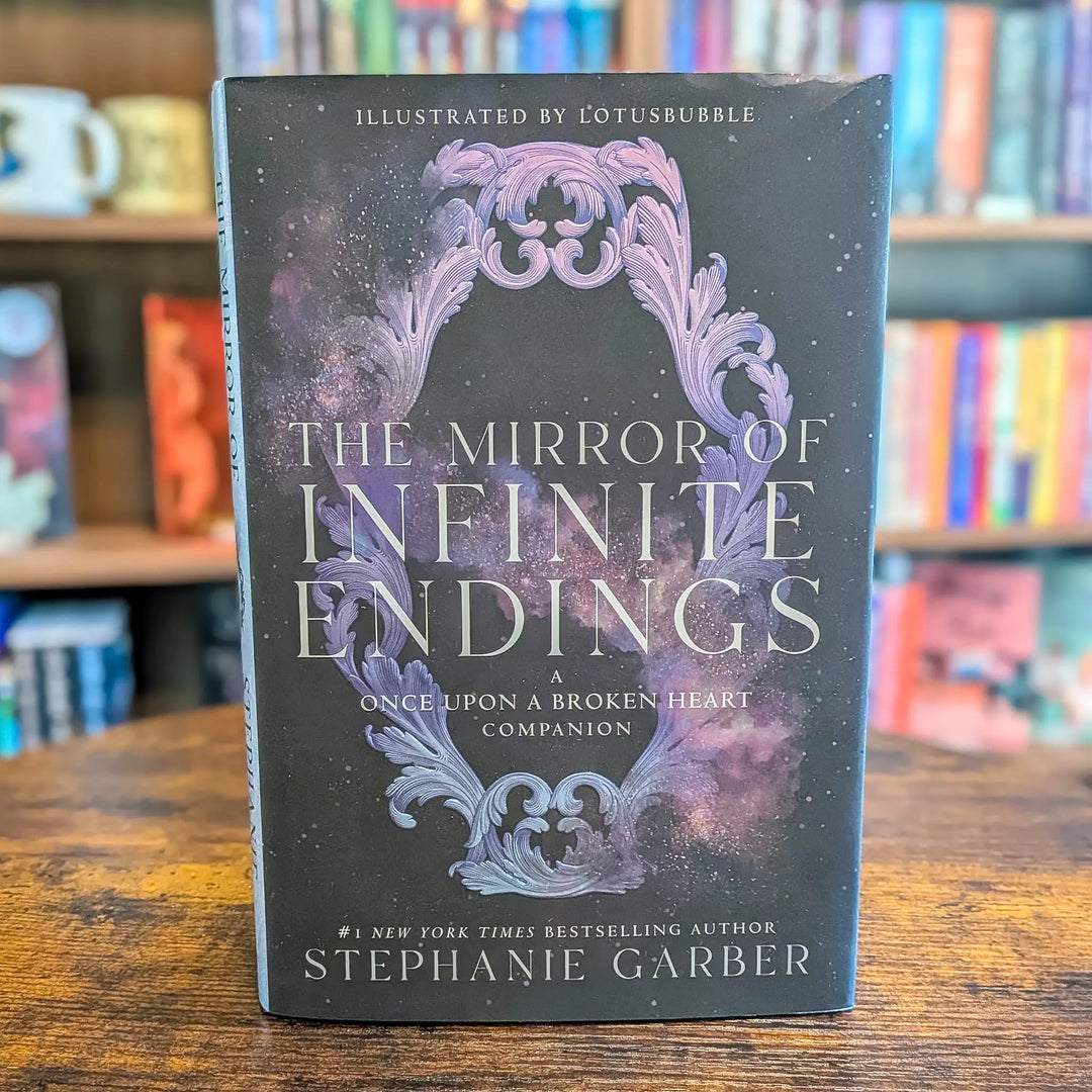 The Mirror of Infinite Endings companion book by Stephanie Garber, standing upright on a wooden table with blurred bookshelves in the background.