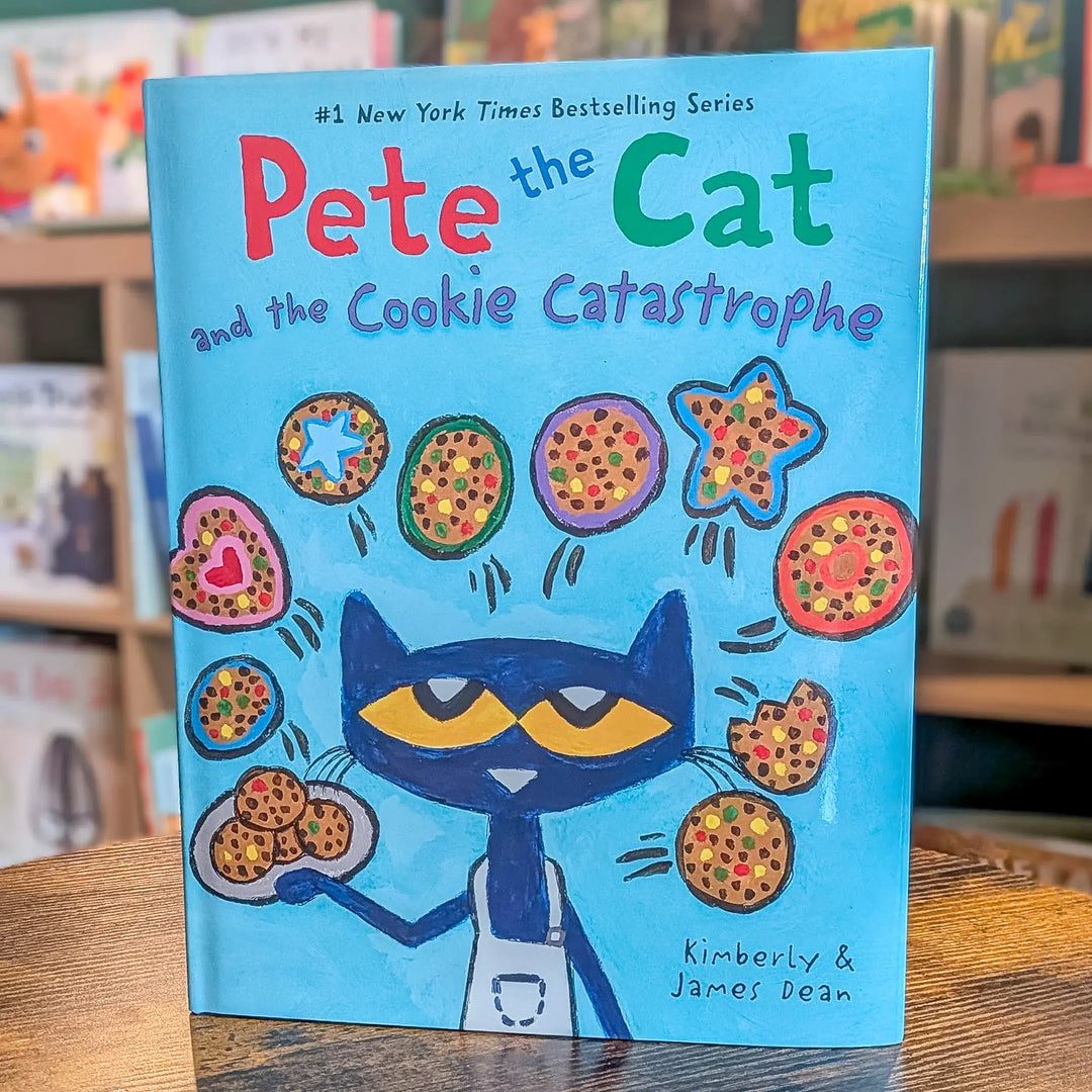 The book 'Pete the Cat and the Cookie Catastrophe' by Kimberly & James Dean, featuring a bright blue cover with Pete the Cat holding cookies, displayed on a wooden table with a blurred bookstore background.