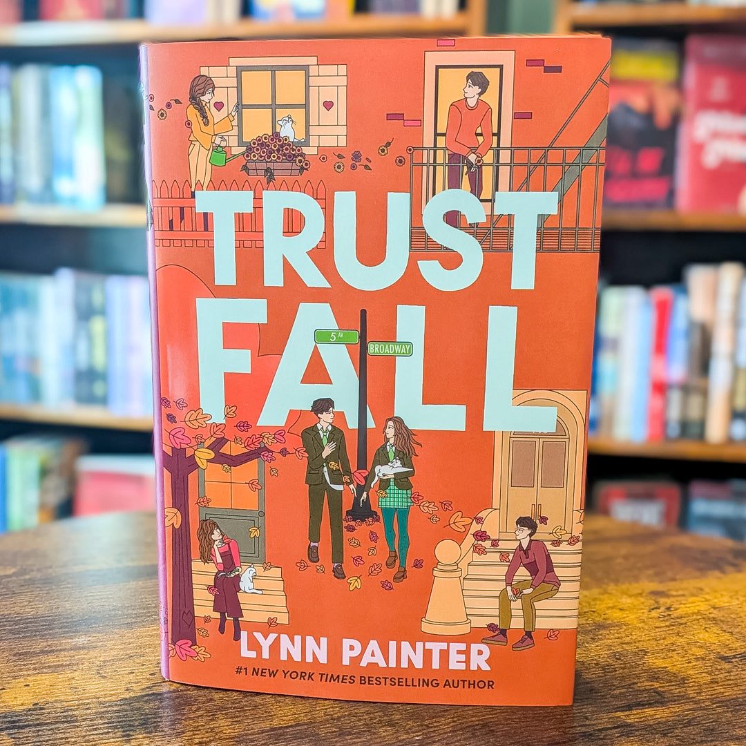 The book 'Trust Fall' by Lynn Painter stands upright on a wooden table with blurred bookshelves in the background. The cover is predominantly orange with large light blue title text and multiple colorful illustrated characters and autumn-themed details.