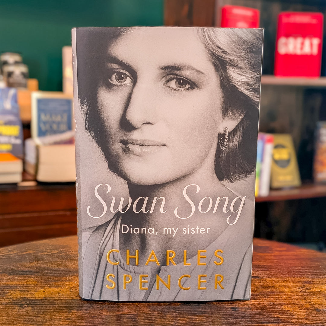 The book 'Swan Song: Diana, my sister' by Charles Spencer stands upright on a wooden table. The cover features a black and white portrait of Princess Diana, with the title in white script and the author's name in gold.