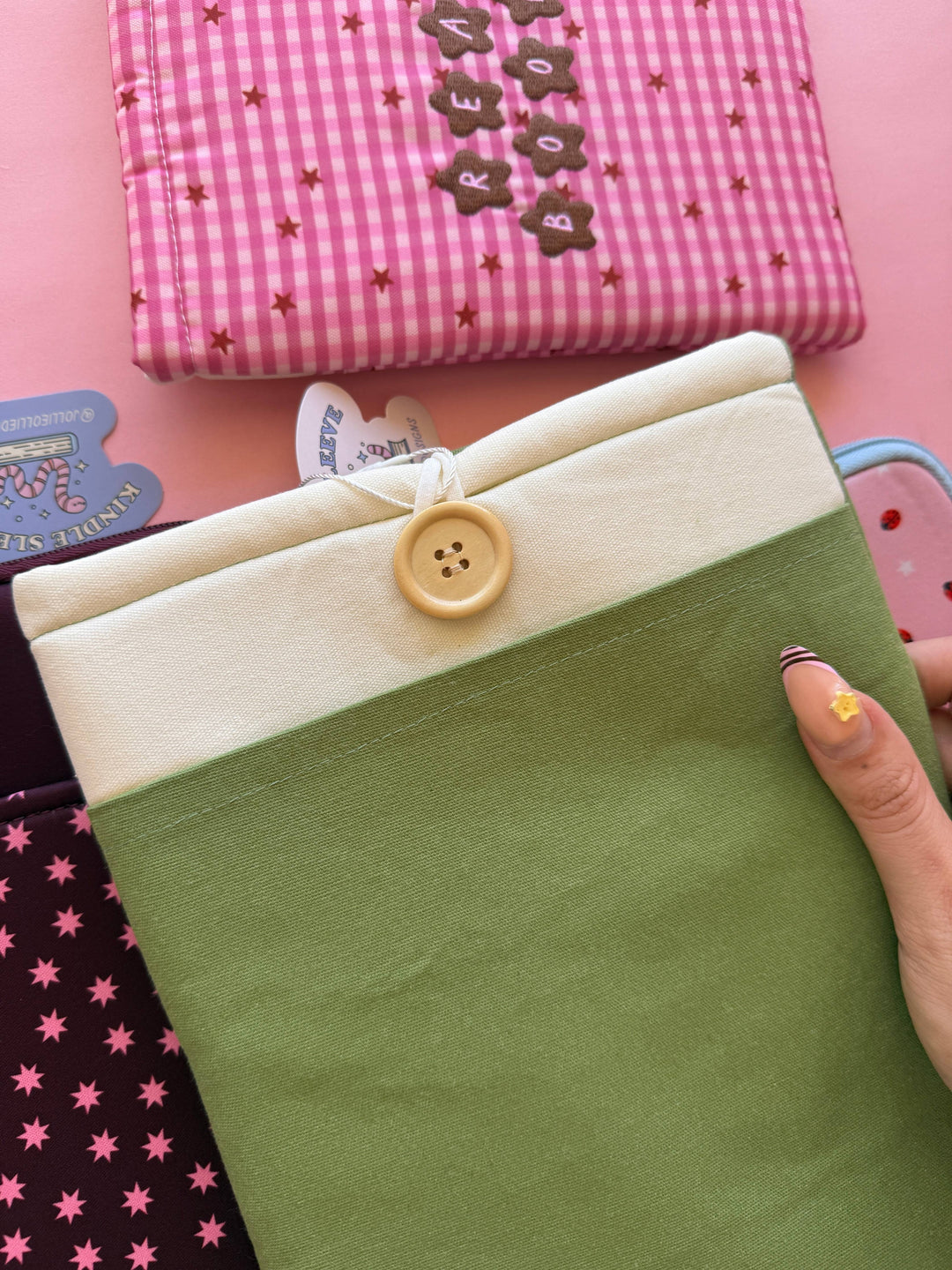 Hand holding a green fabric item with a button, surrounded by other colorful fabric items on a pink background.