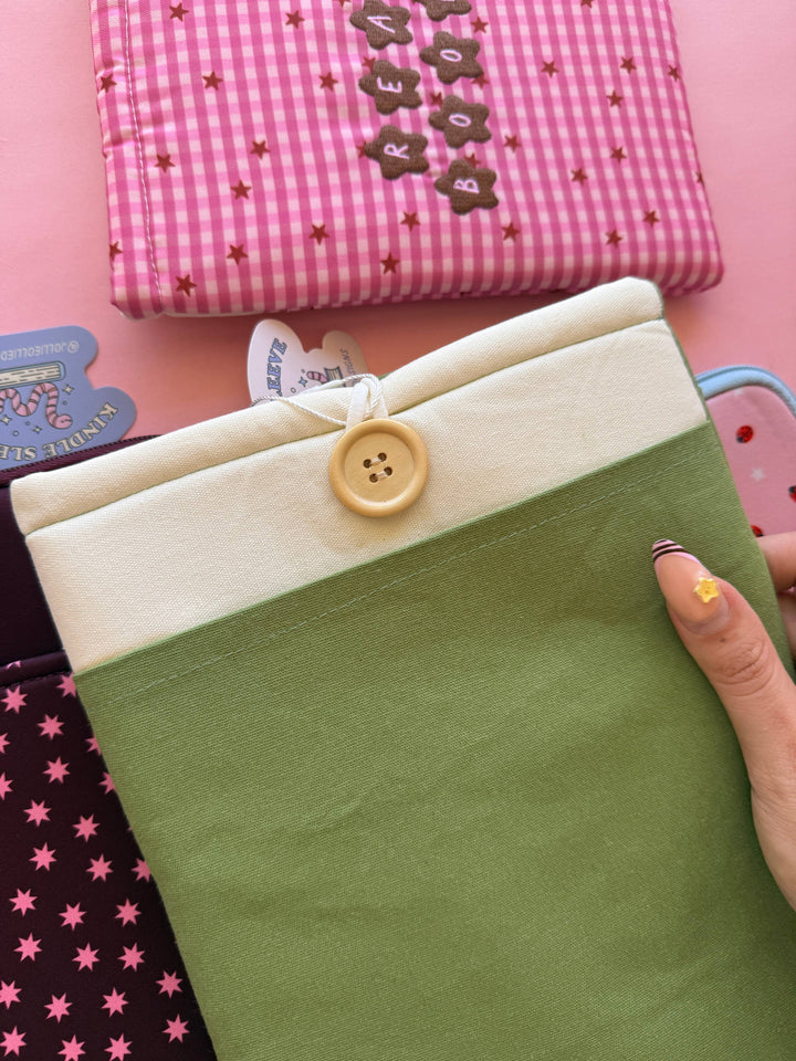 Hand holding a green fabric item with a button, surrounded by other colorful fabric items on a pink background.