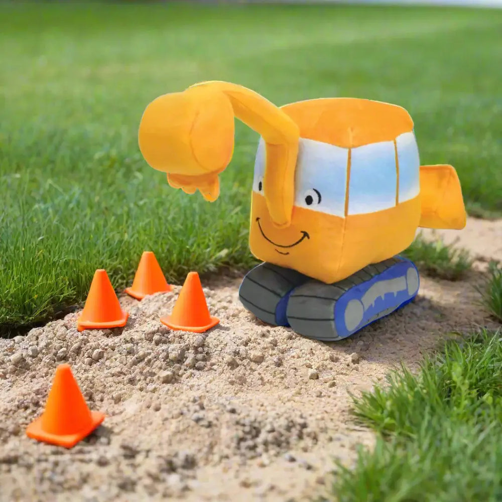 Plush toy excavator with construction cones on a sandy patch with grass in the background