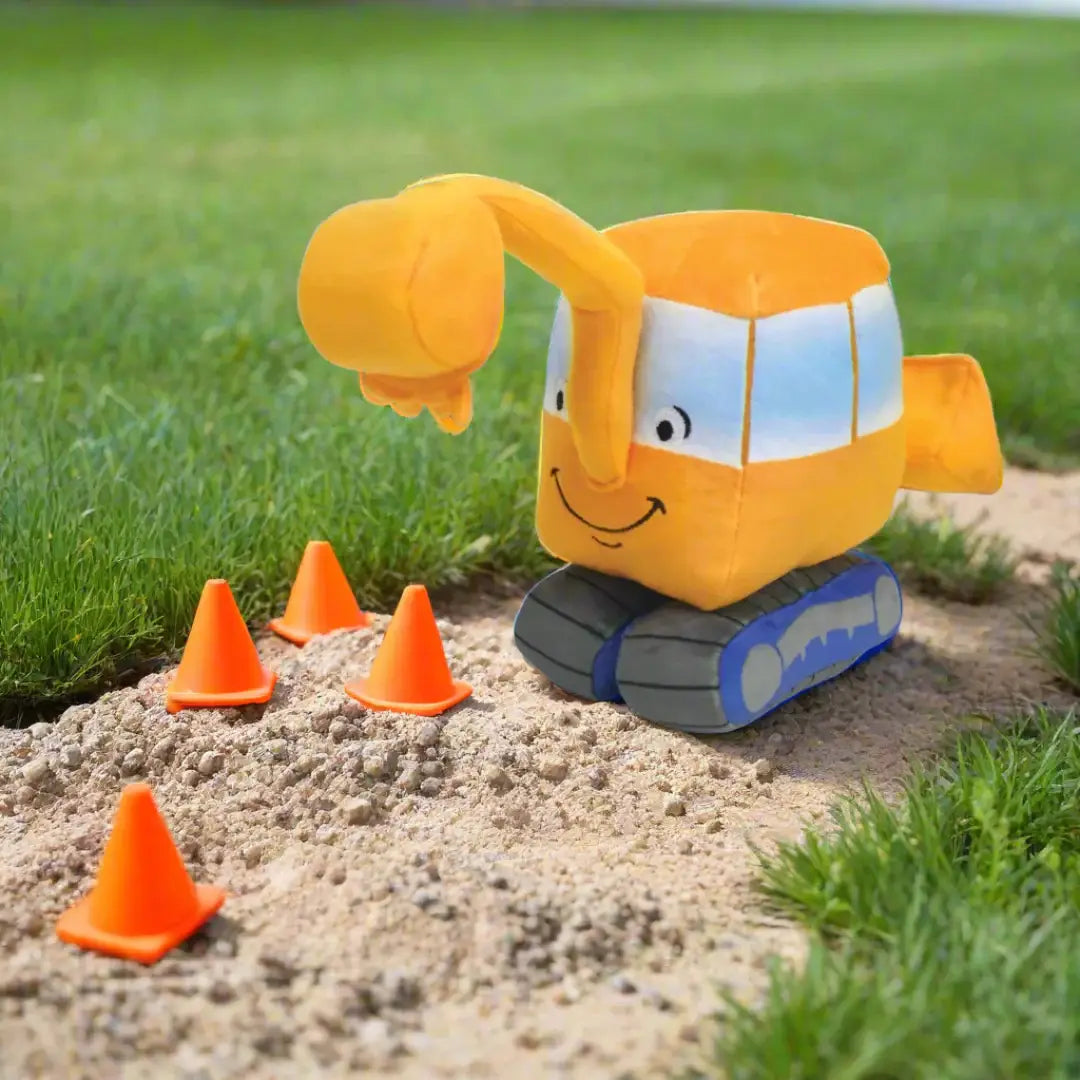 Plush toy excavator with construction cones on a sandy patch with grass in the background