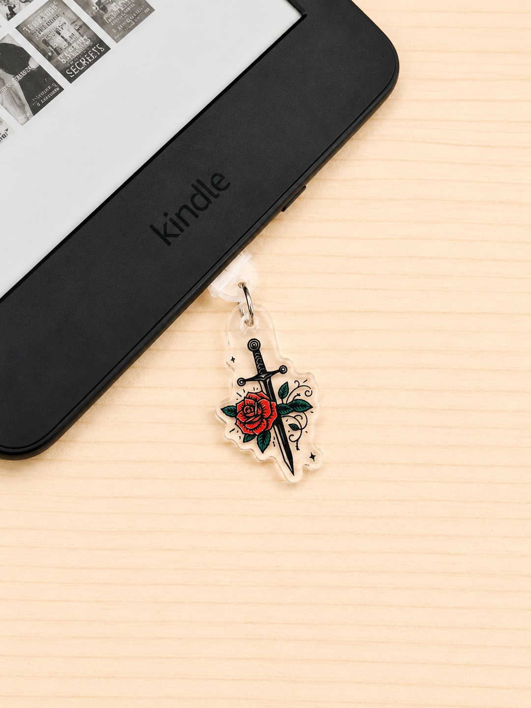 A clear acrylic charm featuring a black dagger and red rose design, hanging from a white loop attached to a Kindle e-reader on a light wood surface.