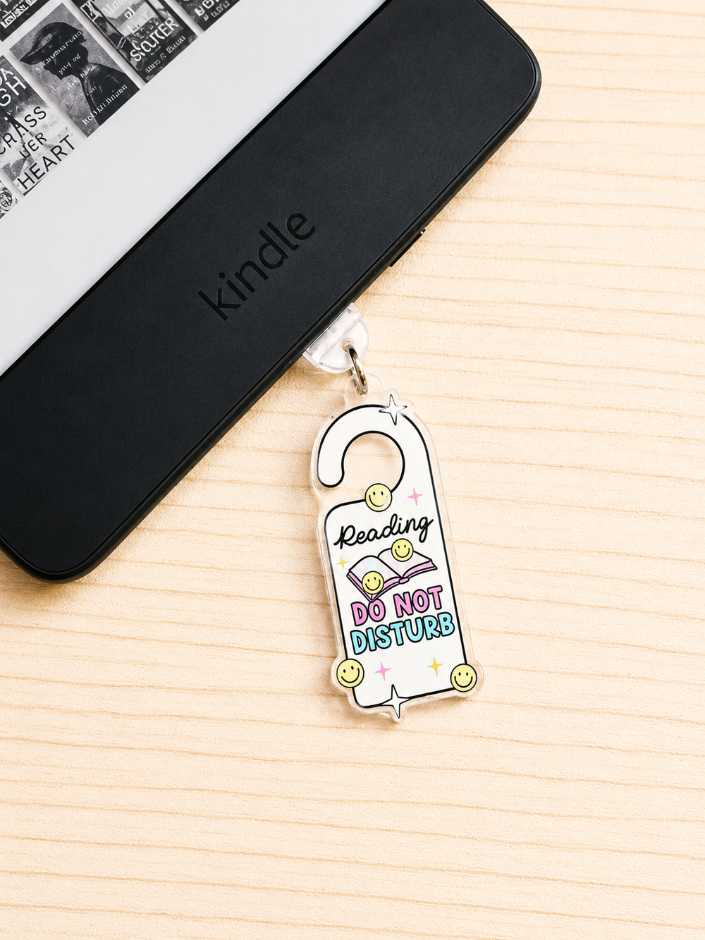 A white enamel library gate hanger charm with colorful text reading 'Reading DO NOT DISTURB' and small smiley faces, shown hanging from a black Kindle device on a light wood surface.